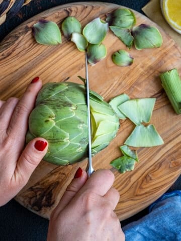 Woman hands cutting off the top of an artichoke on a wooden board.