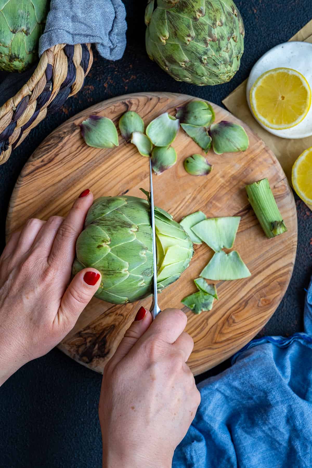 Woman hands cutting off the top of an artichoke on a wooden board.