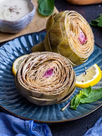 Cooked artichokes on a dark blue plate, a lemon slice and mint leaves on the side.