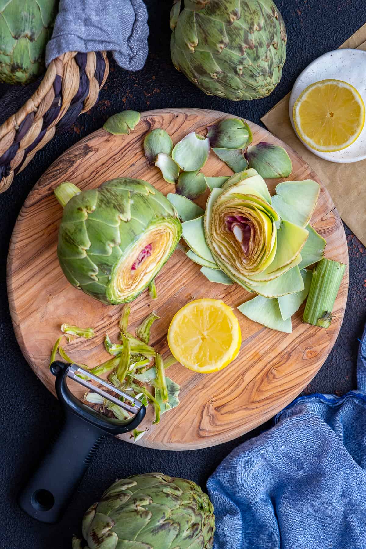 An artichoke trimmed on a wooden board, a slice of lemon and a peeler accompany.