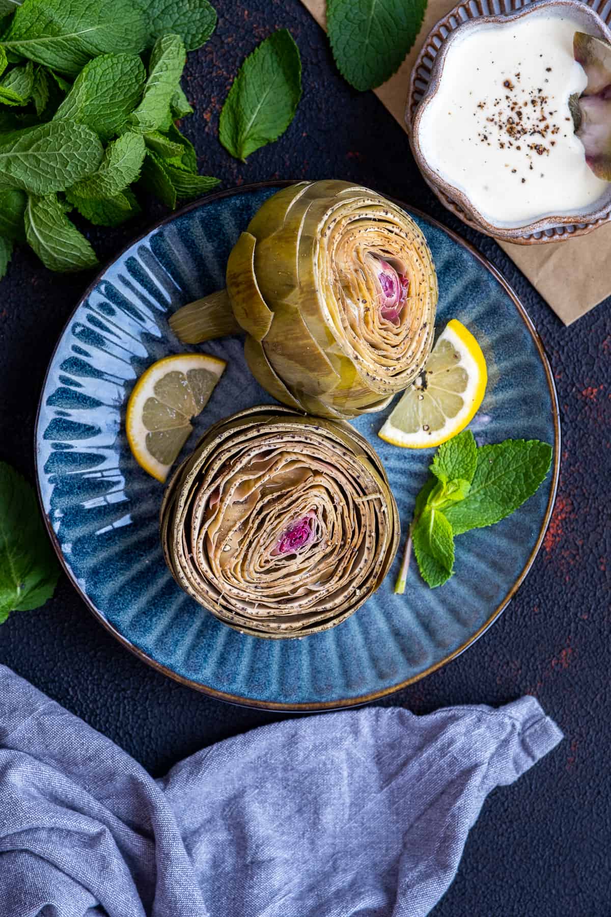 Steamed artichokes garnished with lemon slices and fresh mint leaves on a dark blue plate.