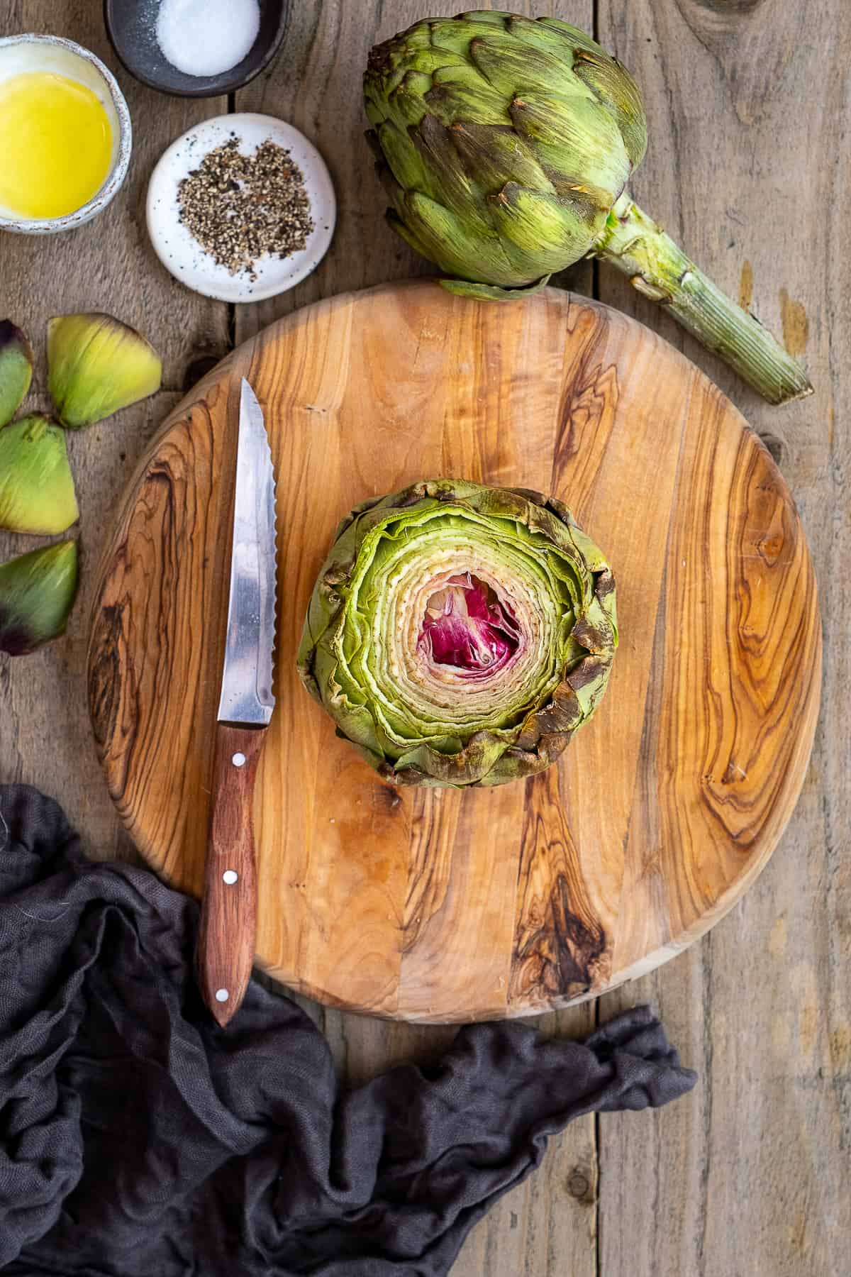An artichoke, top part is cut off placed on a wooden board and a serrated knife on the side.
