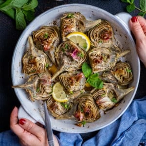 Woman hands holding a round baking pan filled with roasted artichokes garnished with mint leaves and lemon slices.
