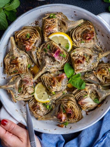 Woman hands holding a round baking pan filled with roasted artichokes garnished with mint leaves and lemon slices.