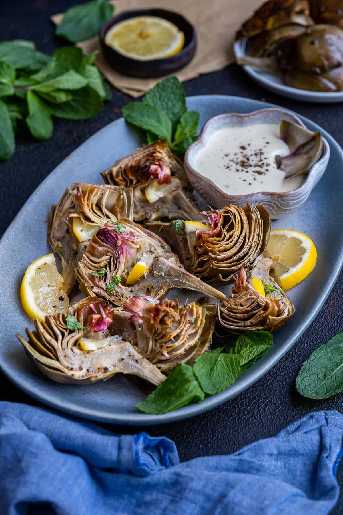Roasted artichokes served on an oval plate with a bowl of dipping sauce on the side photographed from front view.