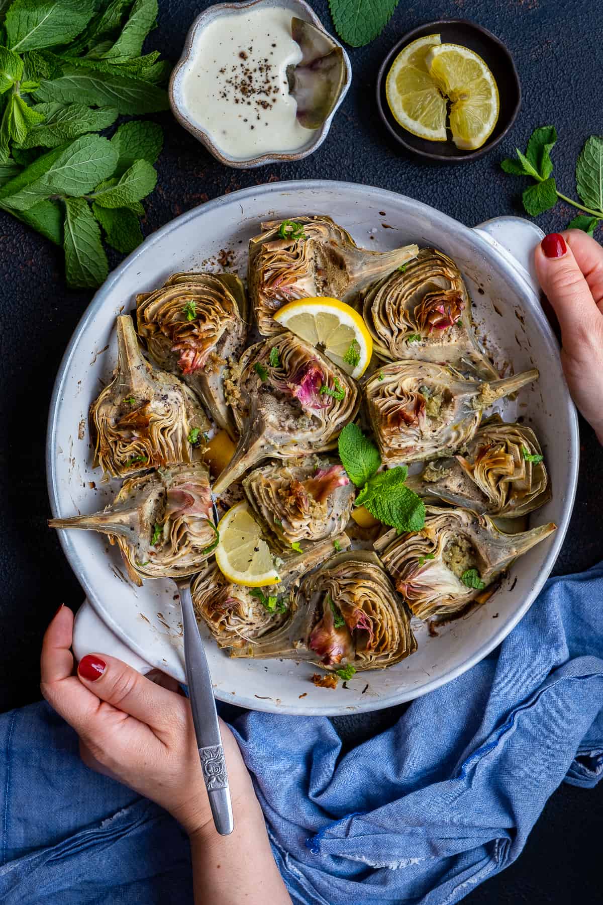 Woman hands holding a white round baking dish filled with roasted artichokes garnished with lemon slices and mint leaves.