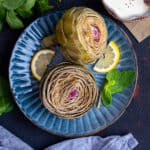 Steamed artichokes garnished with lemon slices and fresh mint leaves on a dark blue plate.