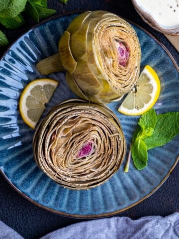 Steamed artichokes garnished with lemon slices and fresh mint leaves on a dark blue plate.