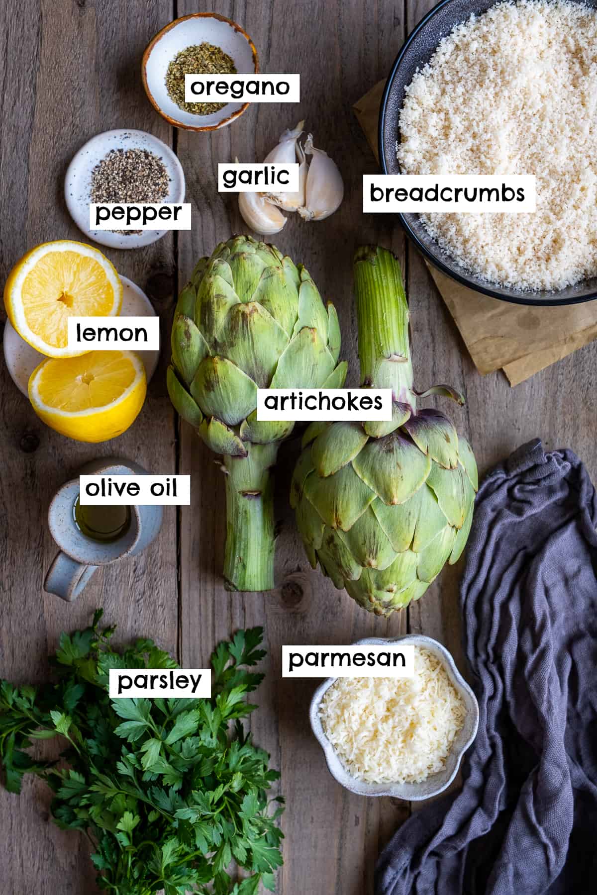 Artichokes, lemon, salt and pepper, breadcrumbs, parmesan cheese, parsley, garlic and olive oil on a wooden backdrop.