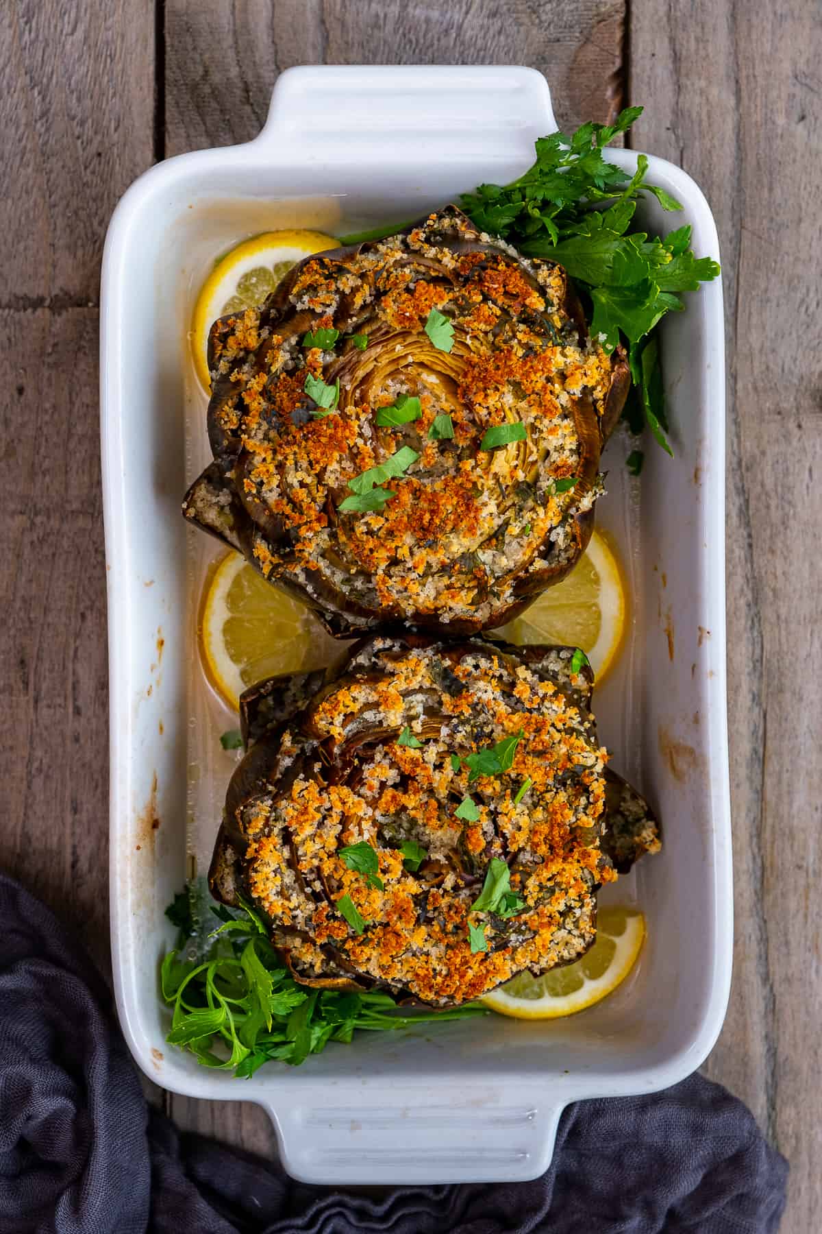 Stuffed artichokes with golden top in a white baking dish, garnished with parsley and lemon slices.