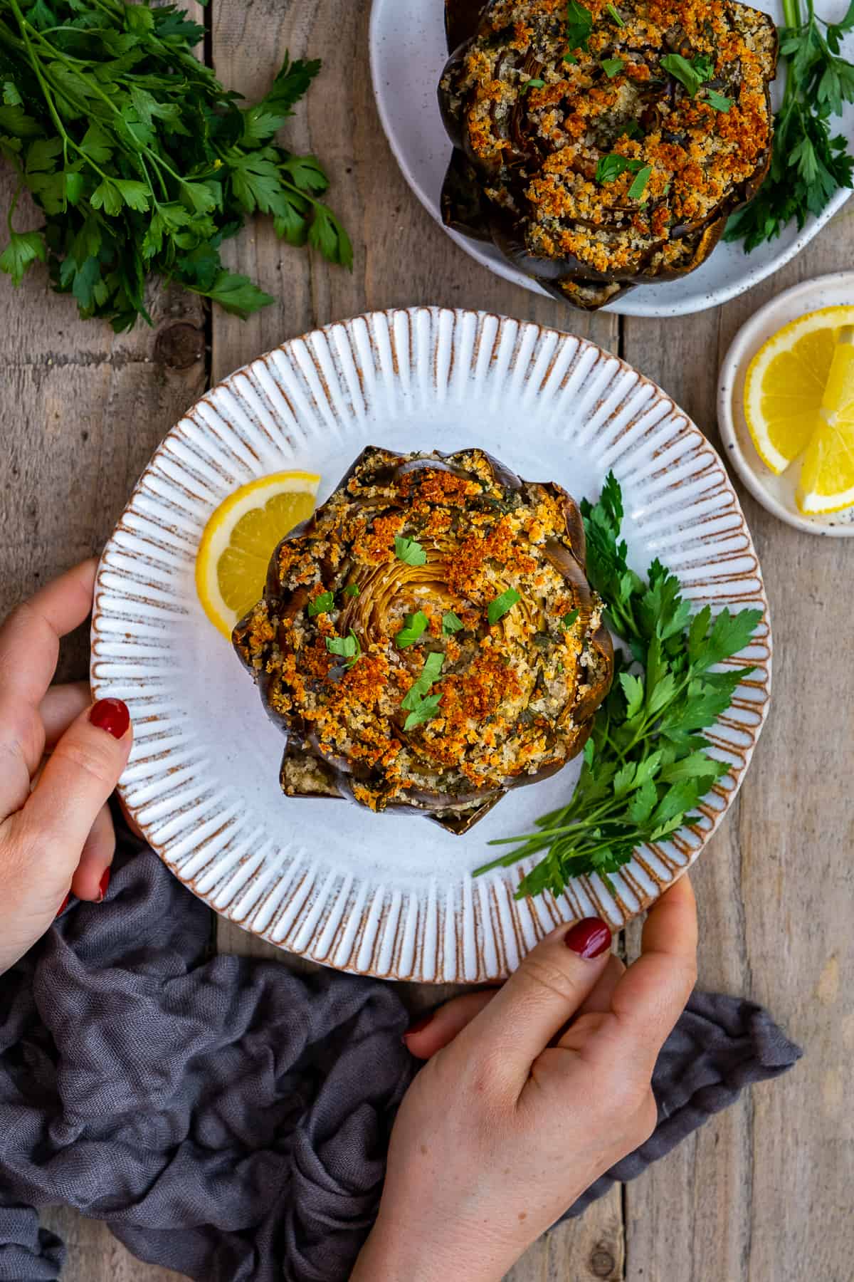 Woman hands holding a plate with a stuffed artichoke garnished with parlsey and a lemon slice.