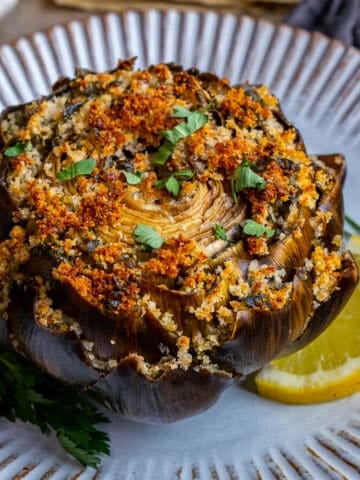 A stuffed artichoke served on a white plate garnished with chopped parsley and a lemon slice.