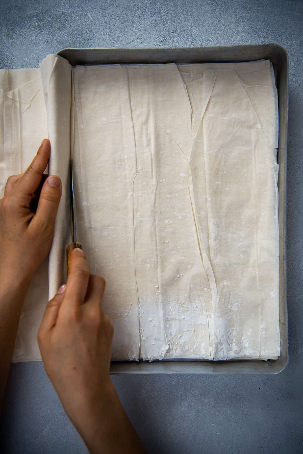 Hands trimming phyllo sheets in a baking pan with a knife.