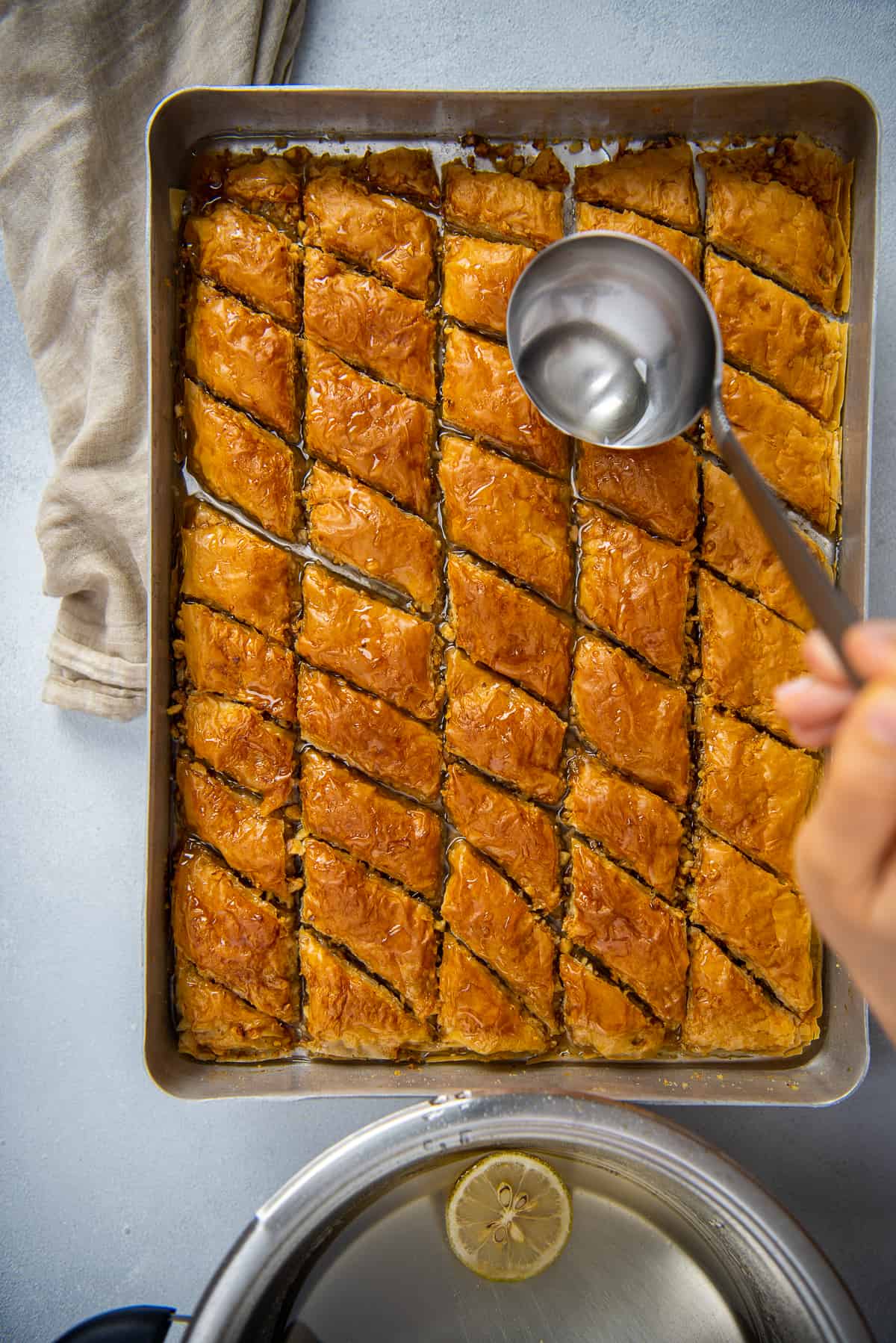 Hands pouring sugar syrup over newly baked Turkish baklava in a baking pan.