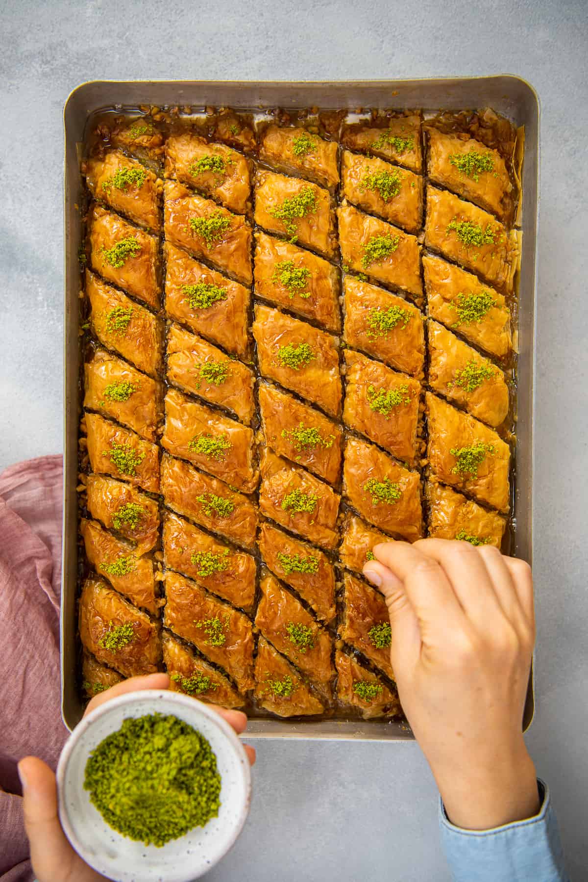 Hands sprinkling ground pistachio over baklava slices in a baking pan.