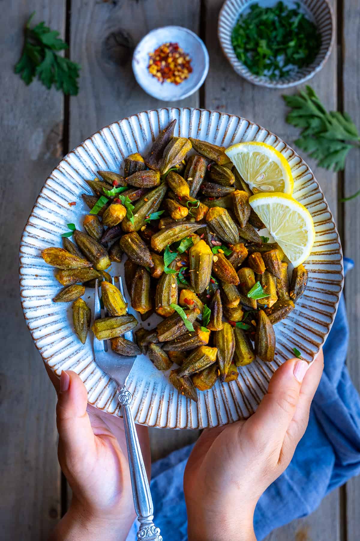 Hands holding a white plate loaded with fried okra garnished with lemon slices and chopped parsley.