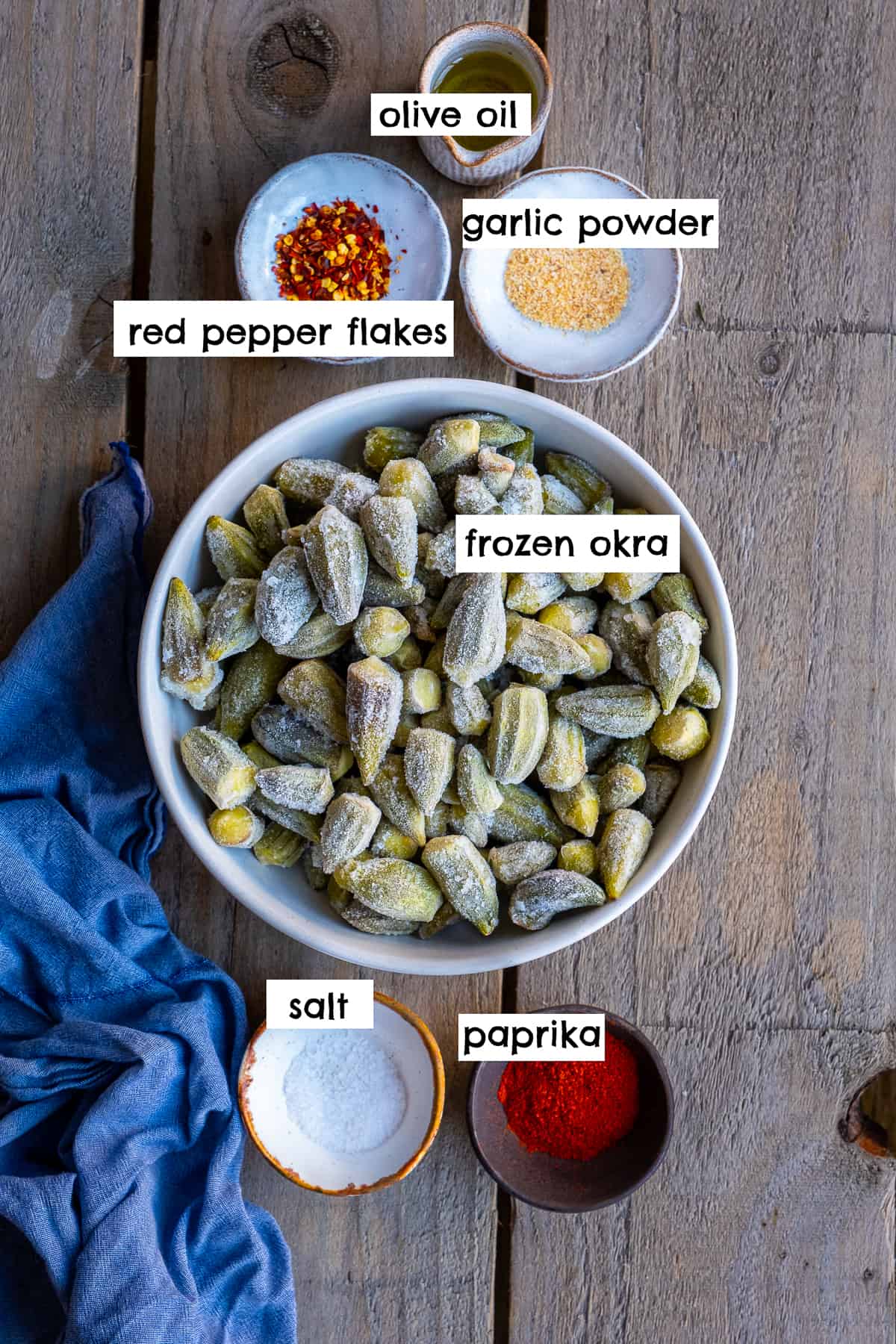 Frozen okra in a bowl, red pepper flakes, olive oil, garlic powder, salt and paprika in small ceramic bowls on a wooden backdrop.