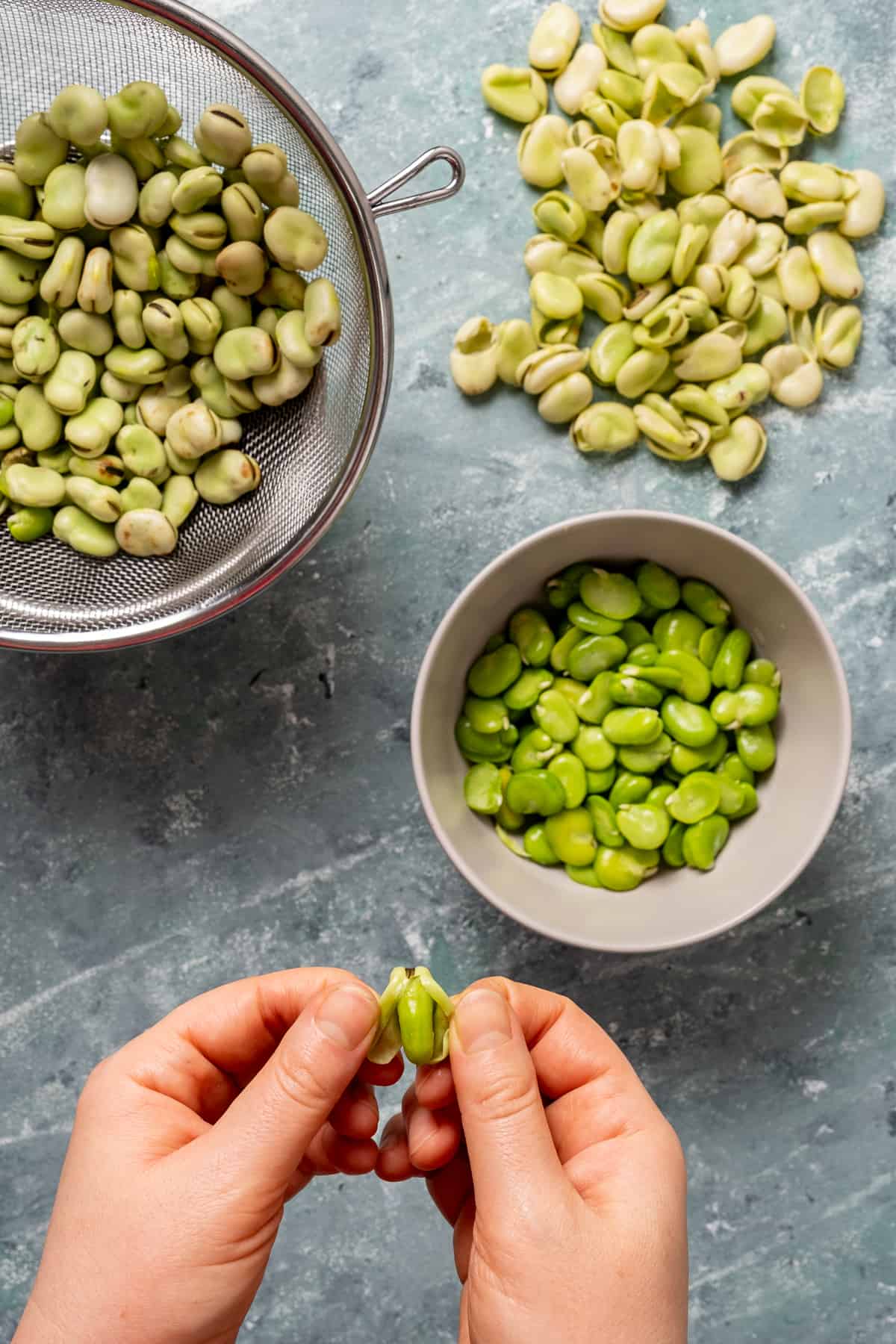 Hands shelling fava beans, shelled fava beans in a bowl, beans with their skin on in a strainer.