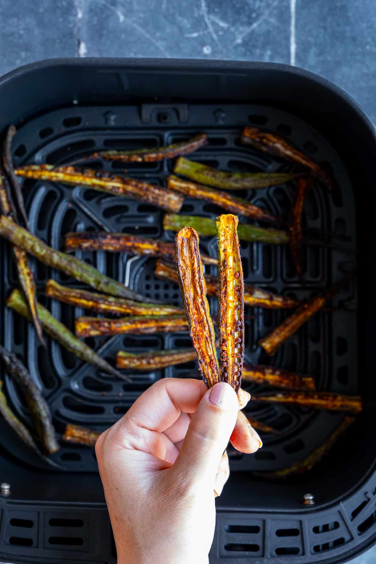 A hand holding two slices of crispy okra fries over an air fryer basket.