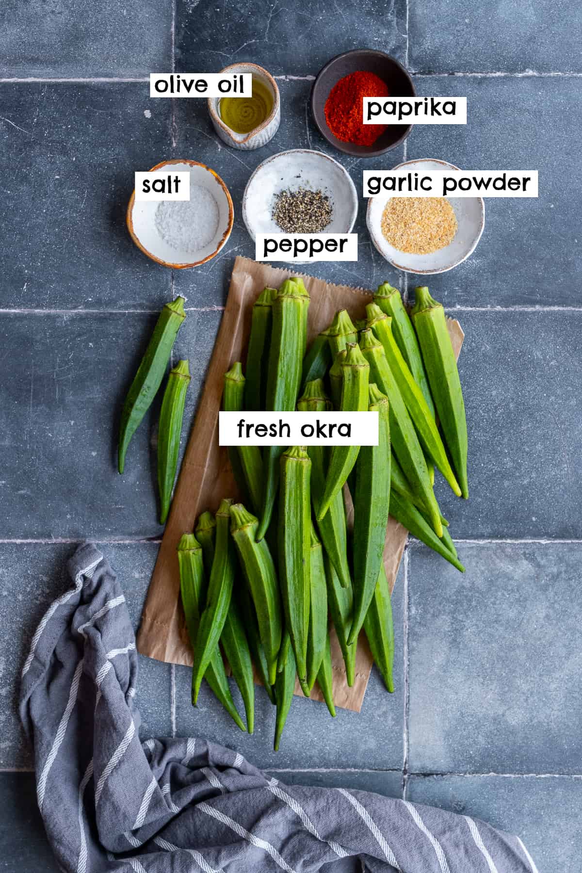 Fresh okra on a piece of brown paper, olive oil, paprika, salt, pepper and garlic powder on grey tiles.