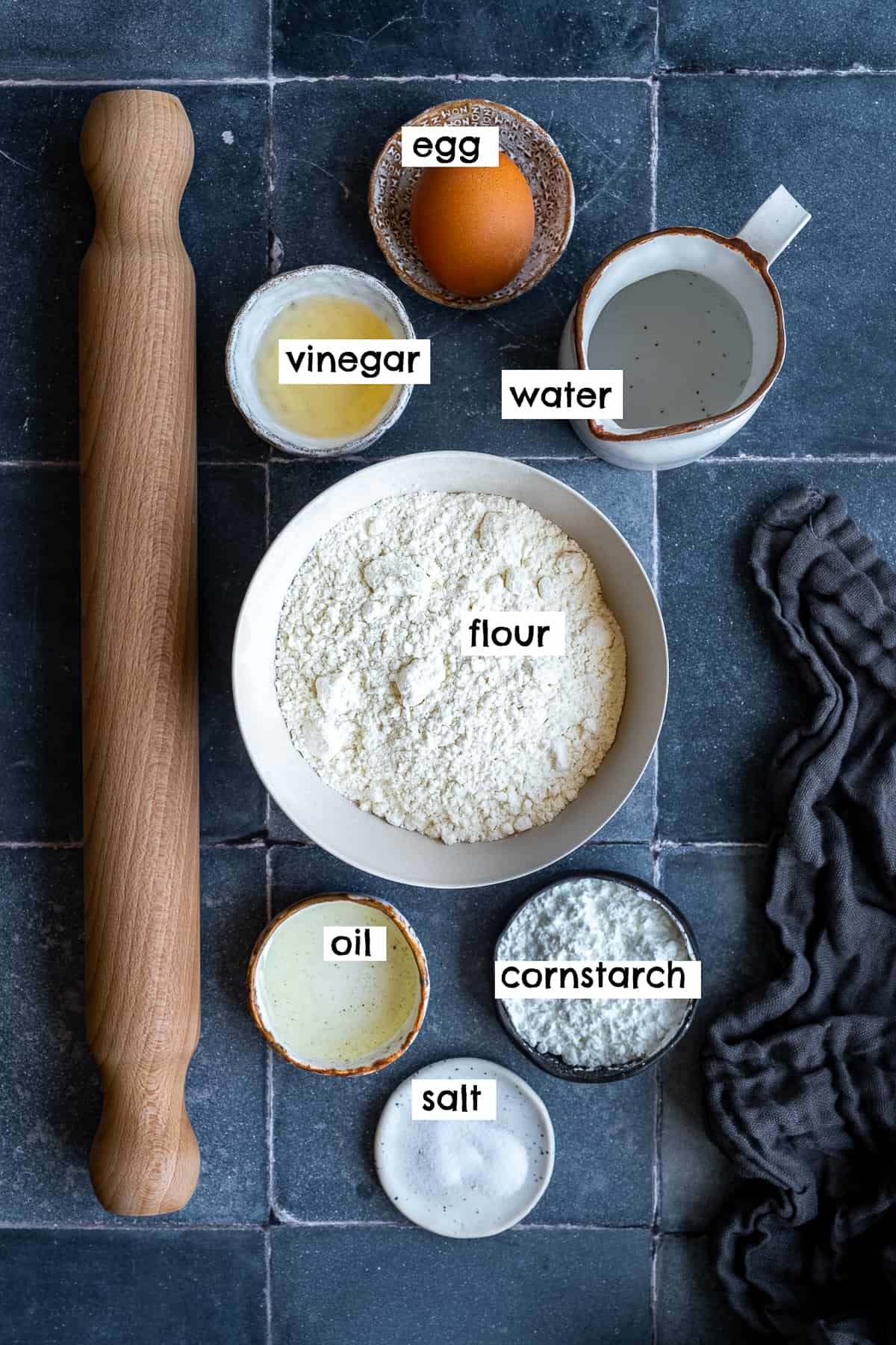 An egg, some flour, cornstarch, salt, oil, vinegar, water and a rolling pin pictured on a grey background.