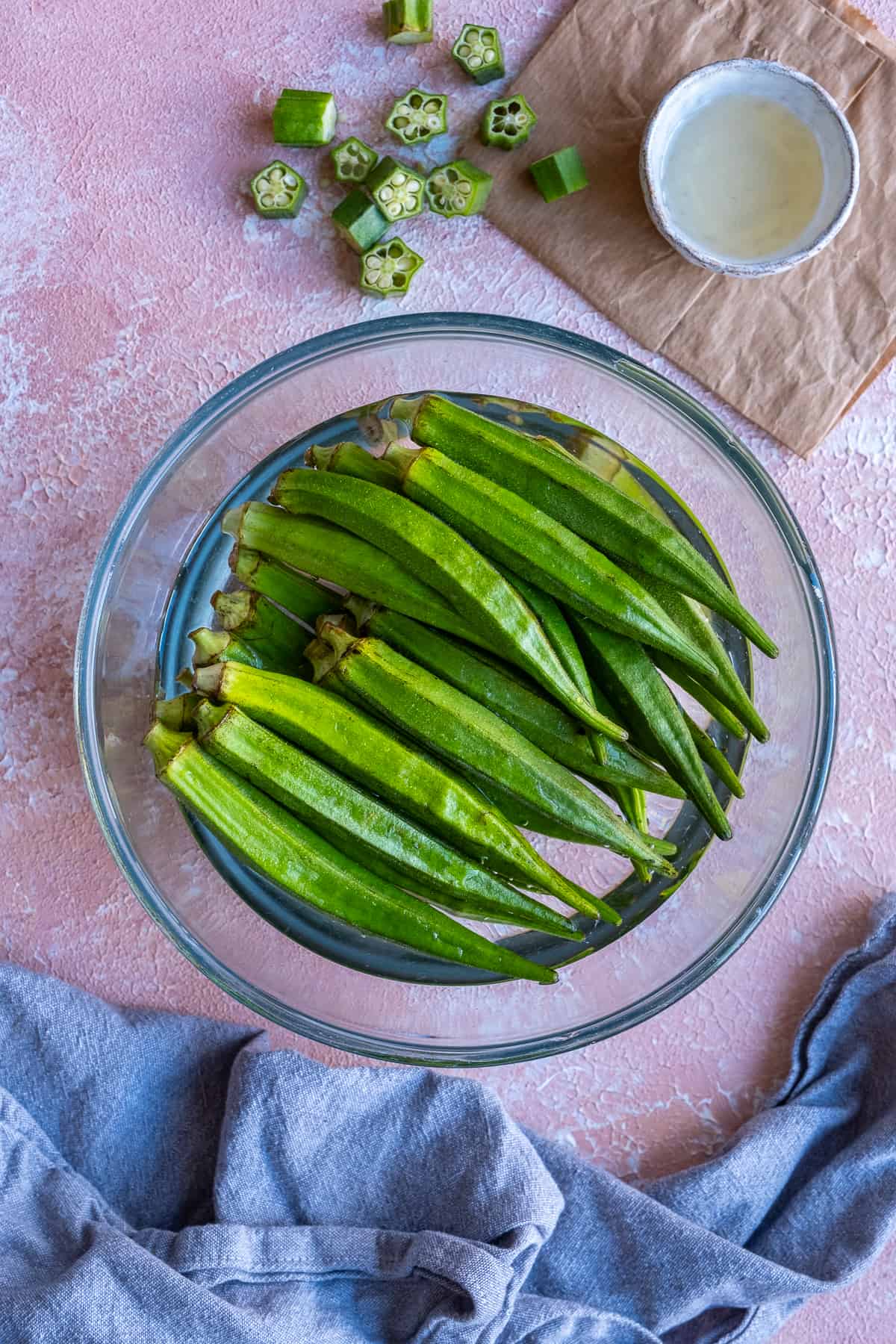 Green okra pods soaked in water in a large glass bowl and vinegar in a small ceramic bowl on the side.