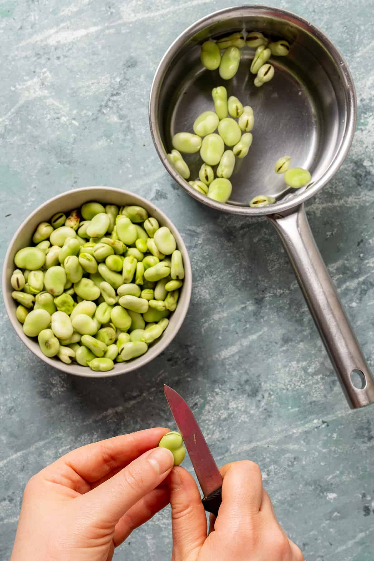 Hands making a slit on fava beans with a knife, fava beans in a bowl and in a saucepan filled with water.