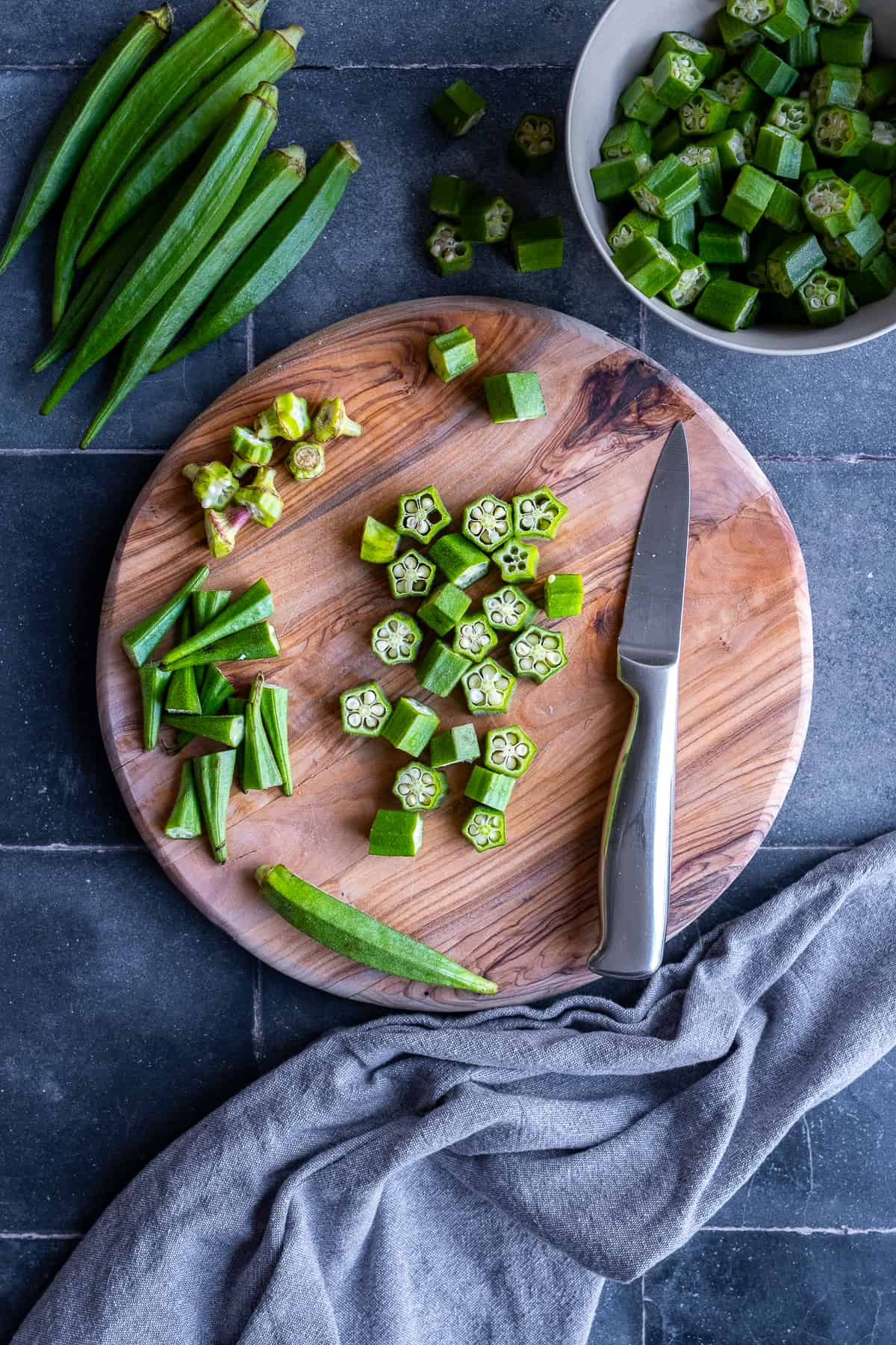 Okra being cut on a round wooden cutting board and a knife on it.