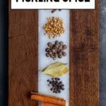 Coriander seeds, mustard seeds, whole cloves, allspice berries, bay leaves, black peppercorns, cinnamon sticks and red pepper flakes placed separately on a wooden cutting board.