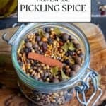 Whole spices like coriander seeds, mustard seeds, allspice berries, cloves, black peppercorns, cinnamon sticks, crushed bay leaves and red pepper flakes combined in a glass jar and pickled okra behind it.