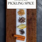 Coriander seeds, mustard seeds, whole cloves, allspice berries, bay leaves, black peppercorns, cinnamon sticks and red pepper flakes placed separately on a wooden cutting board.