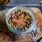 Whole allspice berries, cinnamon sticks, bay leaves, coriander seeds, black peppercorns in a glass jar photographed on a wooden cutting board.