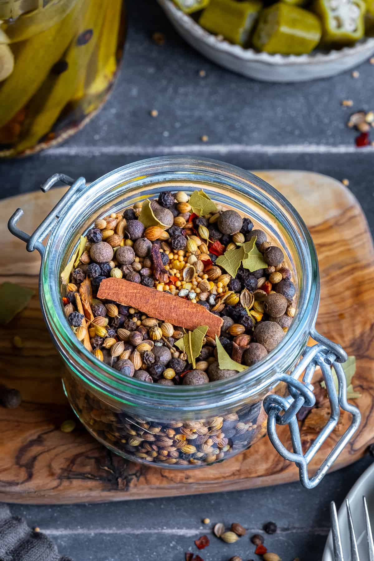 Whole spices like coriander seeds, mustard seeds, allspice berries, cloves, black peppercorns, cinnamon sticks, crushed bay leaves and red pepper flakes combined in a glass jar and pickled okra behind it.