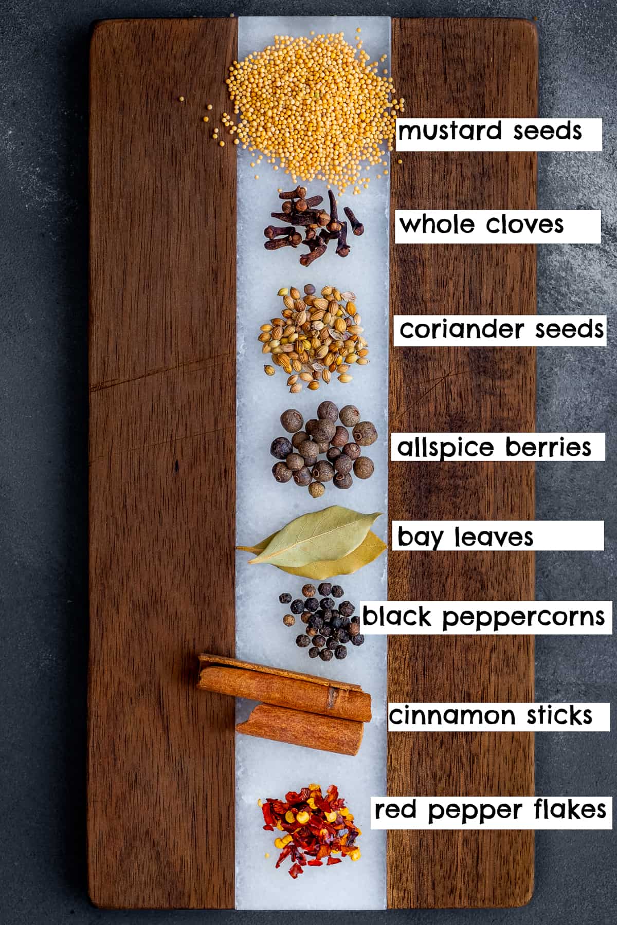 Coriander seeds, mustard seeds, whole cloves, allspice berries, bay leaves, black peppercorns, cinnamon sticks and red pepper flakes placed separately on a wooden cutting board.