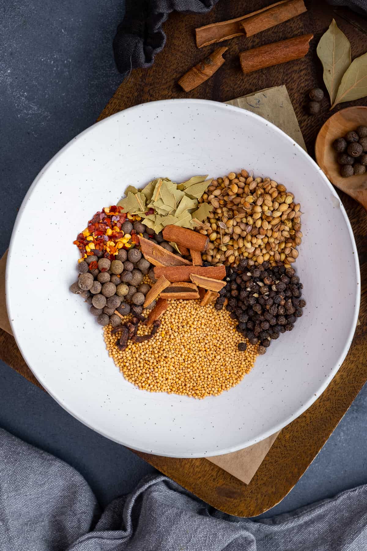 Mustard seeds, allspice berries, coriander seeds, bay leaves, cinnamon sticks and red pepper flakes in a white bowl.