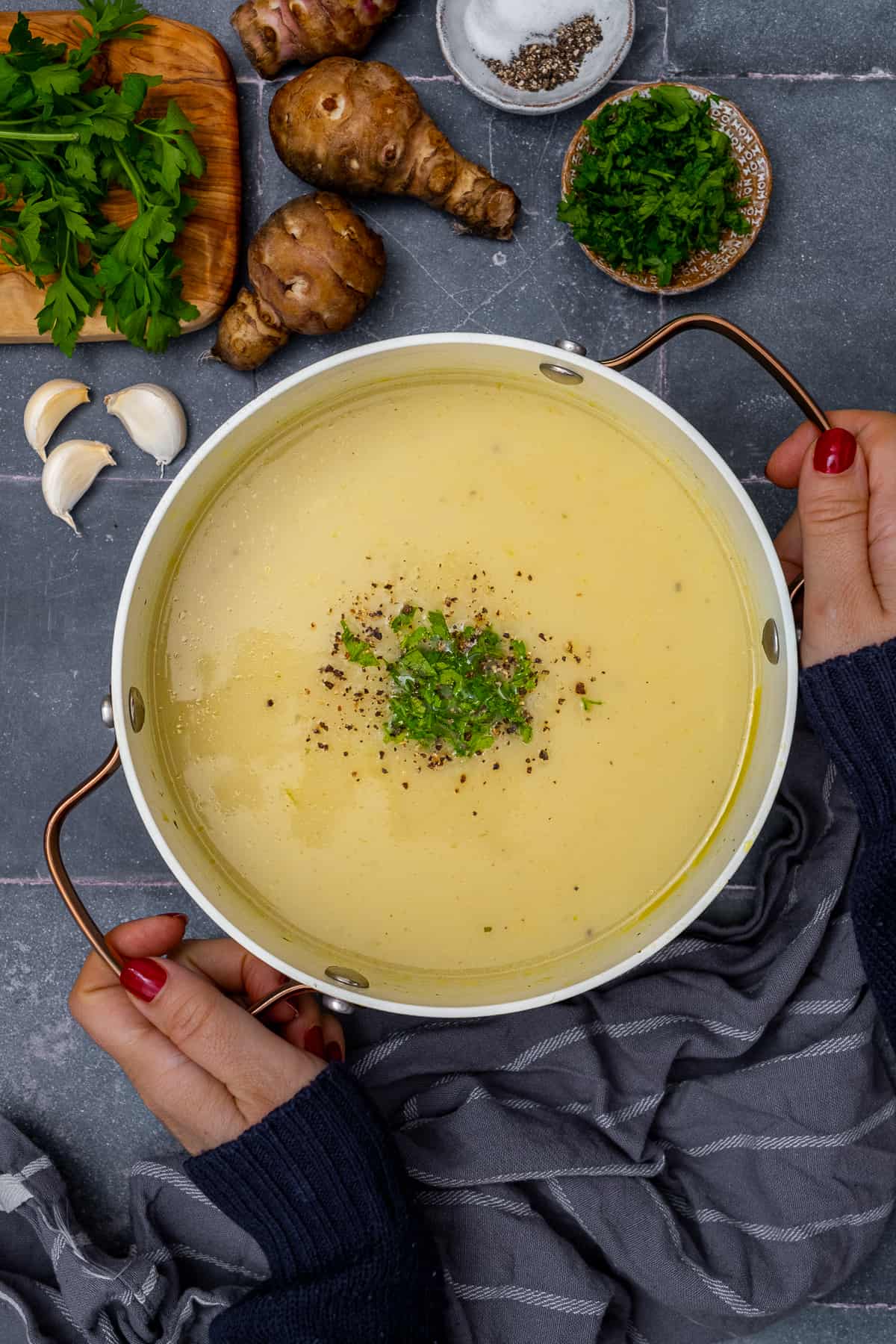 Woman hands holding a pot of Jerusalem artichoke soup garnished with parsley and black pepper.