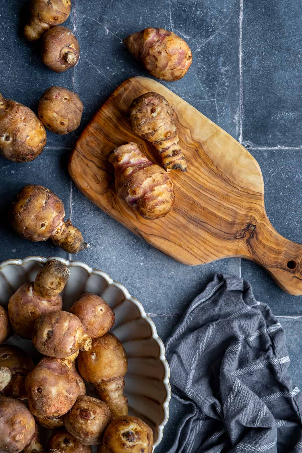 Jerusalem artichokes on a wooden cutting board and in a large bowl photographed on a dark background.