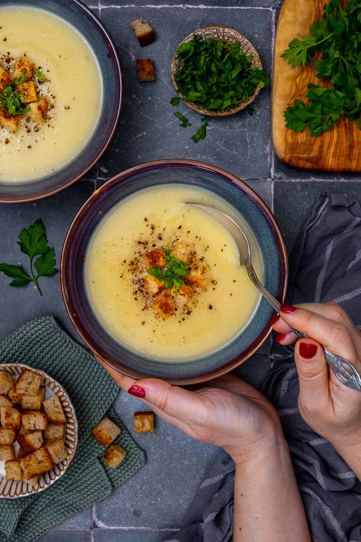 Woman hands holding a bowl of Jerusalem artichoke soup and a spoon, another bowl of soup and croutons on the side.