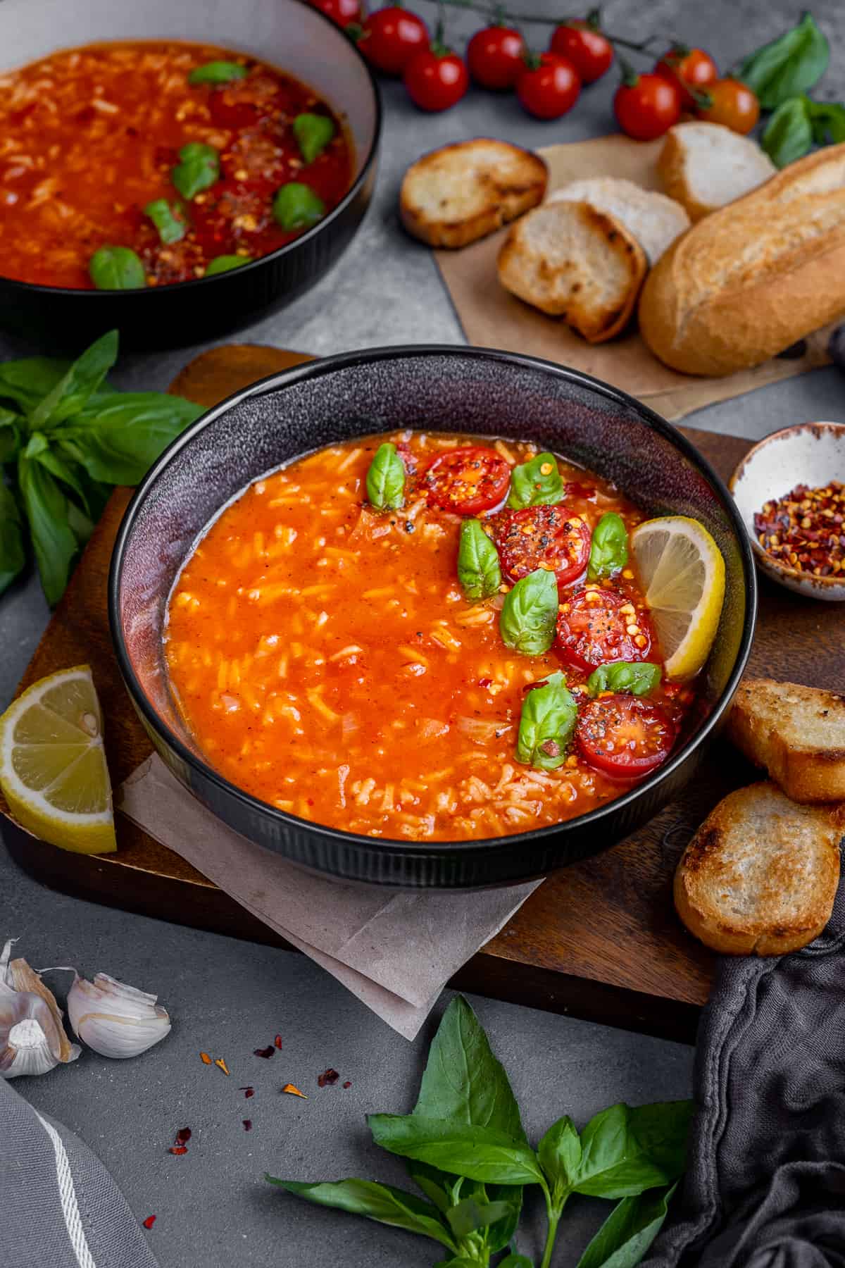 Tomato rice soup served in a black bowl, topped with fresh basil leaves, halved cherry tomatoes and a slice of lemon.