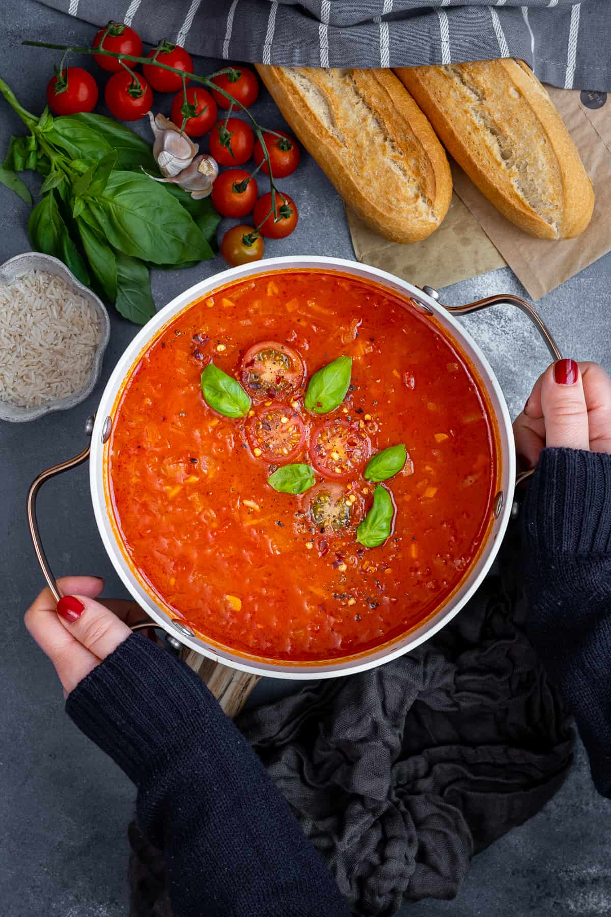 Hands holding a pot of tomato and rice soup garnished with fresh basil leaves and halved cherry tomatoes.