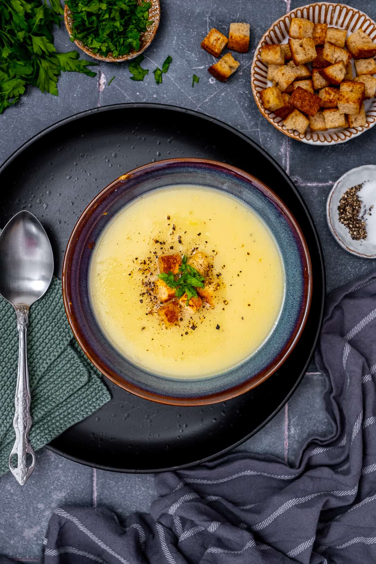 Jerusalem artichoke soup garnished with croutons and parsley placed on a black plate, a spoon on the side.