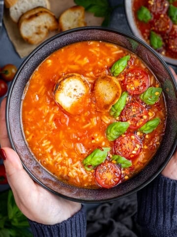 Hands holding a black bowl filled with tomato rice soup garnished with fresh basil and halved cherry tomatoes.