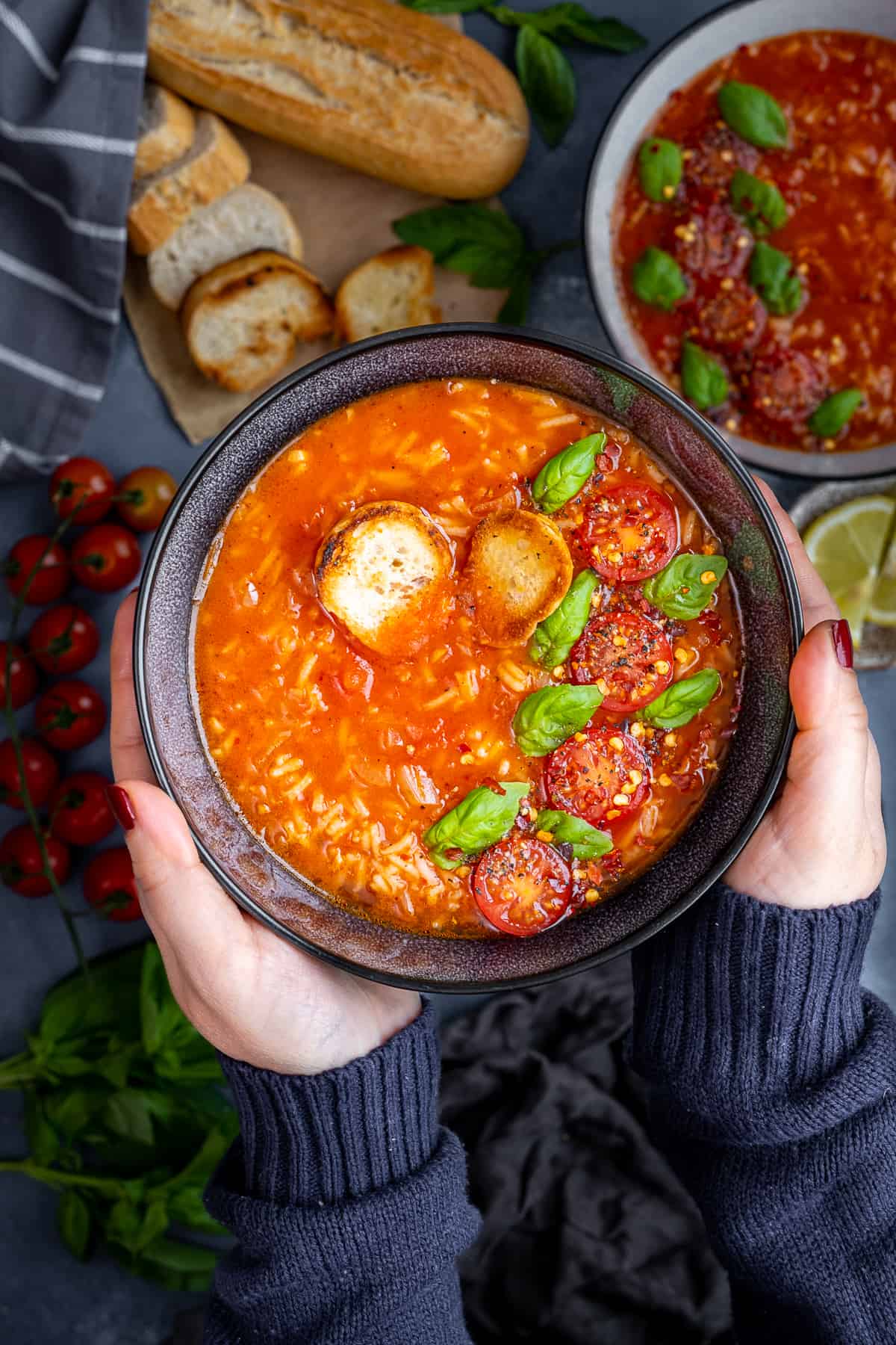 Hands holding a bowl of tomato rice soup topped with fresh basil leaves, halved cherry tomatoes and toasted bread pieces.