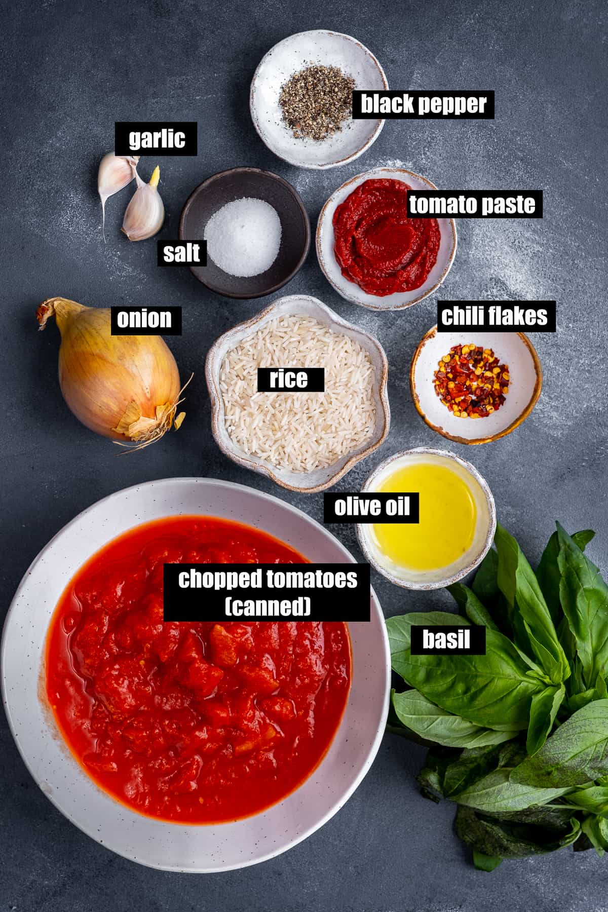 Chopped tomatoes, uncooked rice, tomato paste, red pepper flakes, salt and black pepper, onion, garlic cloves, olive oil and fresh basil on a dark backdrop.