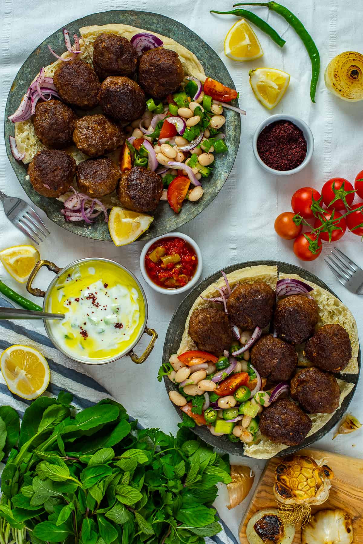 Meatballs served with bean salad on two plates, roasted garlic, a yogurt dip, cherry tomatoes and herbs on the side.