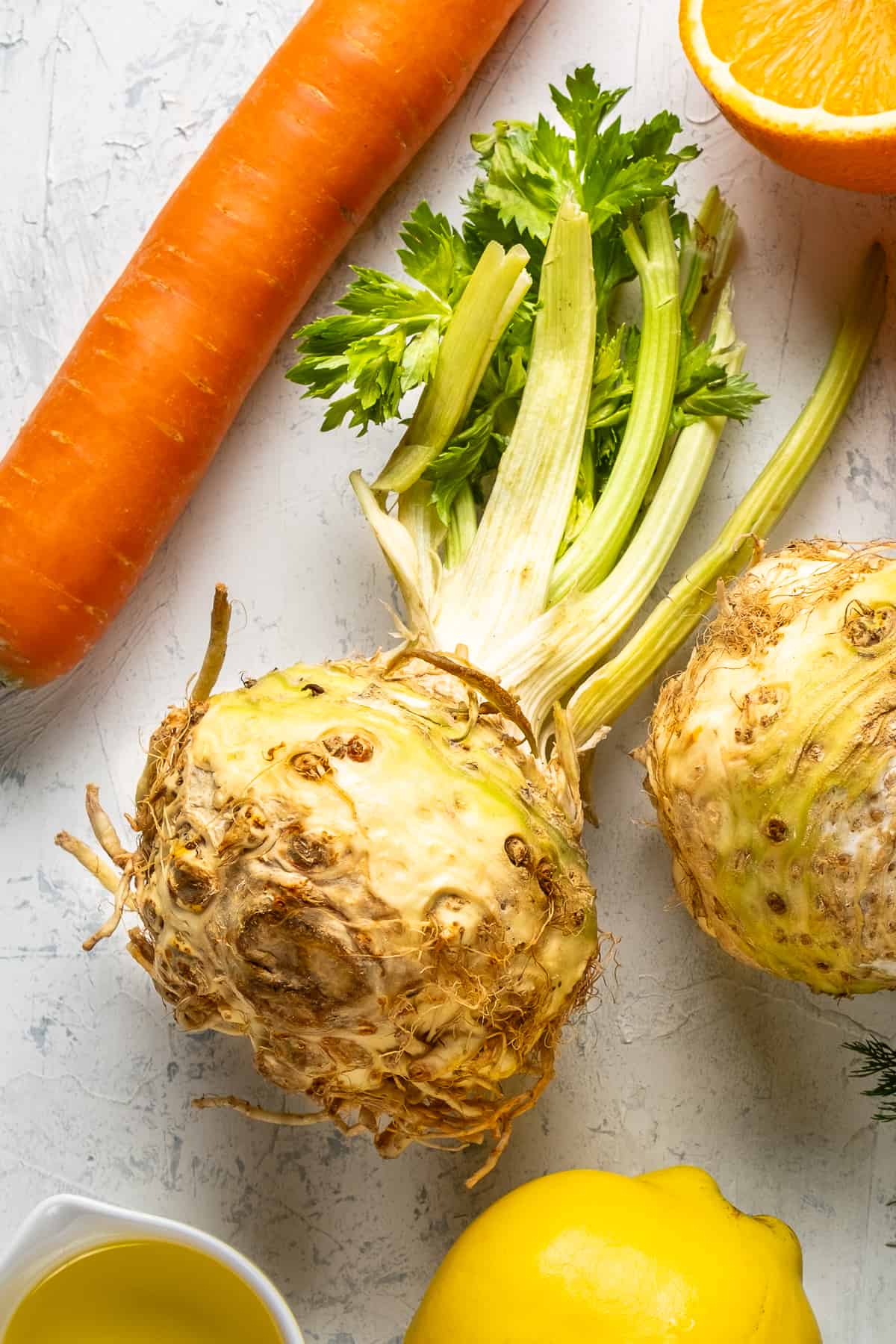 A whole celeriac with its greens pictured with a carrot and orange on a white marble.