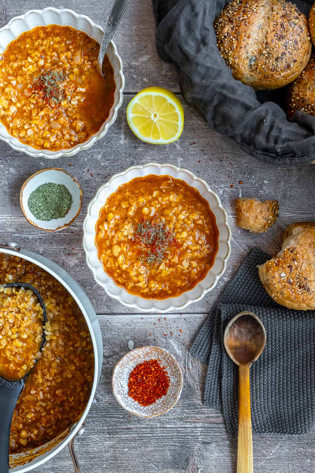 Ezogelin soup in two bowls and in a pot, dried mint, red pepper flakes bread rolls and a wooden spoon accompany.