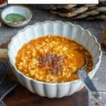 Ezogelin soup topped with red pepper flakes and dried mint in a white bowl, a spoon inside it and extra spices behind it.