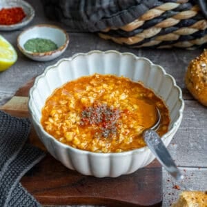 Ezogelin soup topped with red pepper flakes and dried mint in a white bowl, a spoon inside it and extra spices behind it.