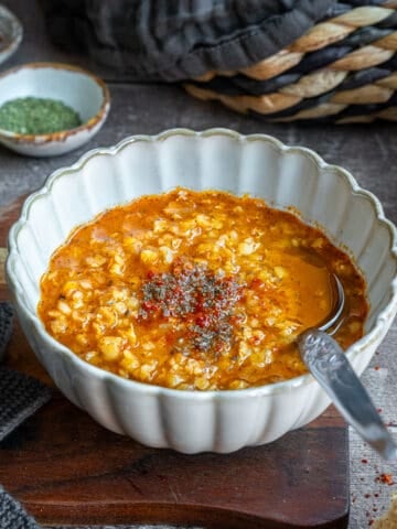 Ezogelin soup topped with red pepper flakes and dried mint in a white bowl, a spoon inside it and extra spices behind it.